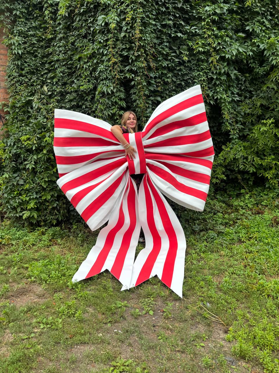 May include: An oversized decorative bow featuring a repeating pattern of bold red and white vertical stripes. The large bow has two wide loops and two long, pointed tails extending downward, constructed from a stiff material to maintain its shape.