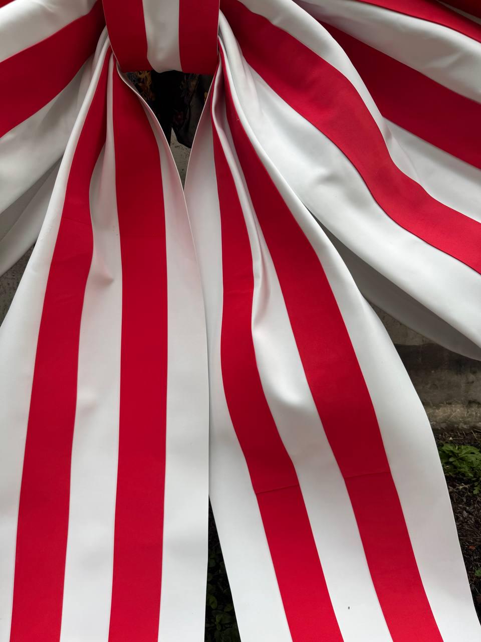 May include: A large decorative bow made of smooth material featuring a repeating pattern of vertical red and white stripes. The bow is tied in a central knot with two long, wide tails hanging downward, displaying the consistent striped design throughout.