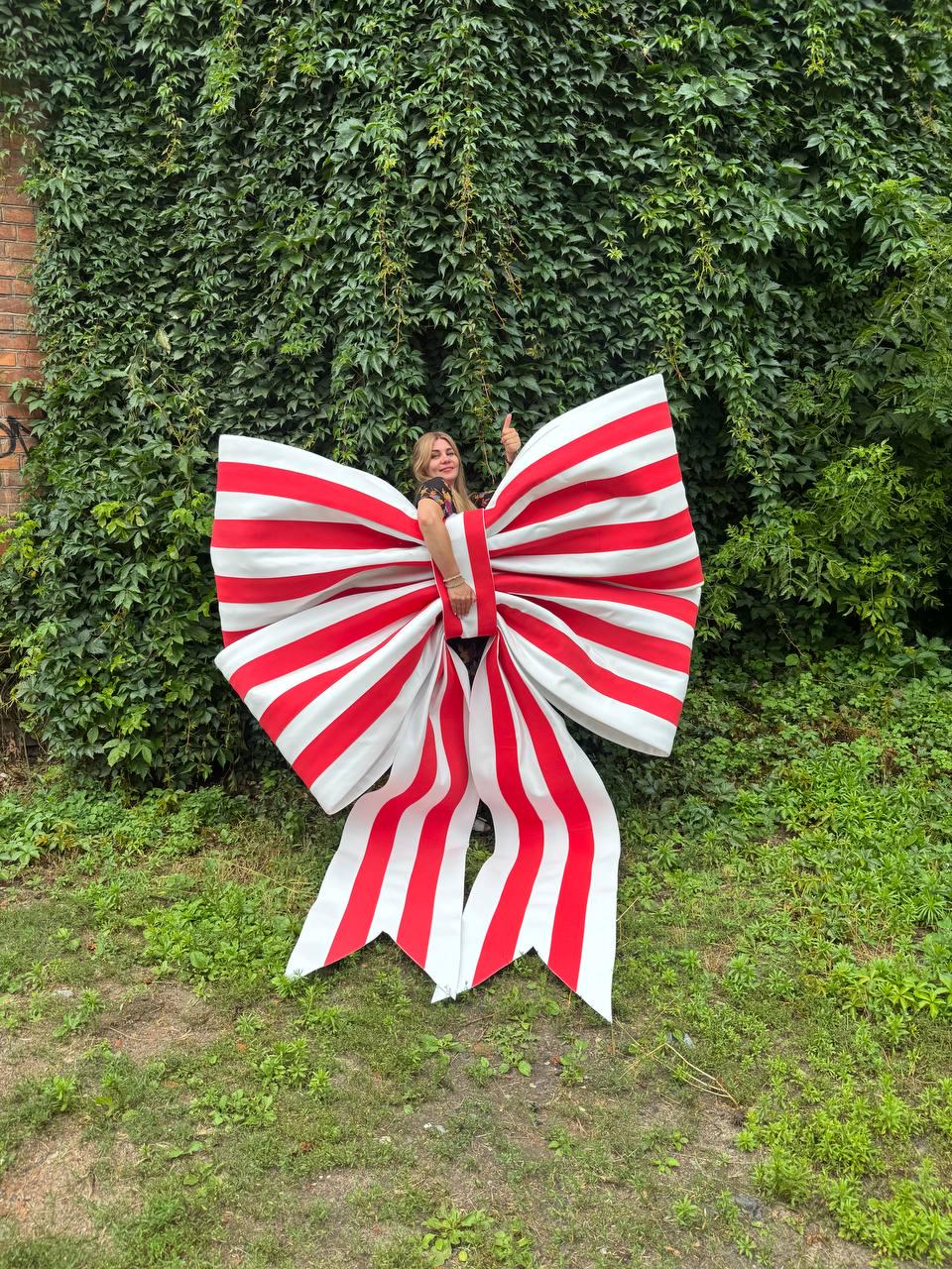 May include: An oversized decorative bow featuring bold vertical red and white stripes. The large fabric accessory is constructed with two wide loops and two long, pointed tails hanging downward, creating a prominent display piece with a classic pattern.