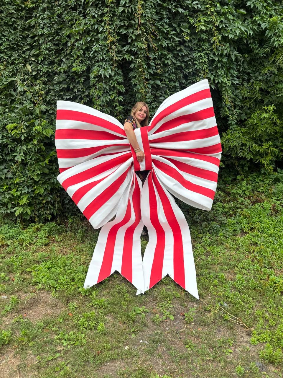 May include: An oversized decorative bow featuring a bold pattern of alternating vertical red and white stripes. The large bow has two wide loops and two long, pointed tails hanging downward, constructed from a stiff material to maintain its shape.