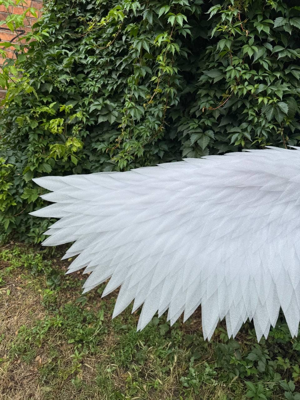 May include: A pair of large, white, feathered-style wings constructed from layered, pointed, semi-translucent material. The wings feature a detailed, overlapping design that mimics the appearance of natural plumage, extending horizontally across the frame.