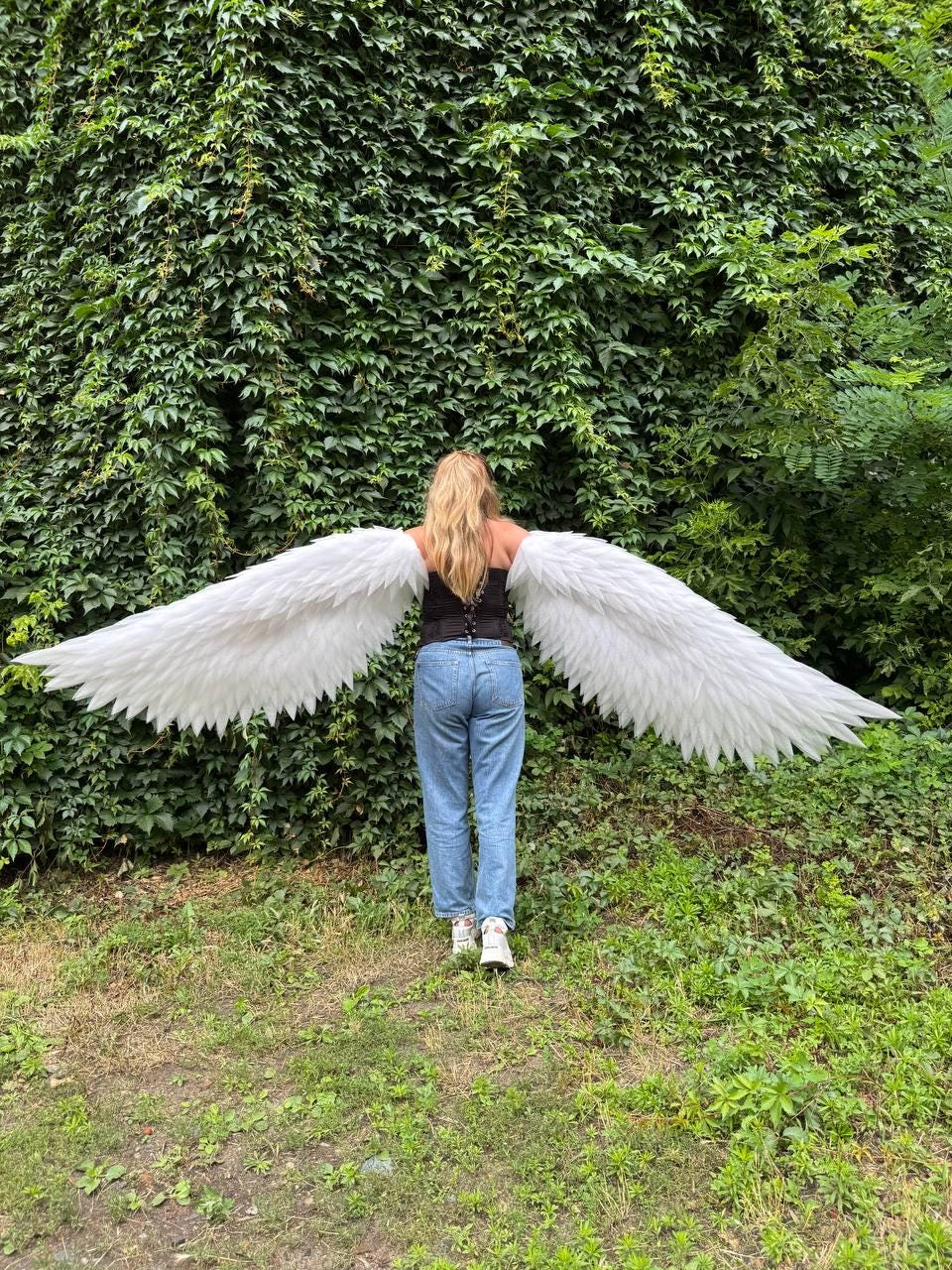 May include: Large, expansive white feathered wings attached to a black corset top worn with light blue denim jeans. The wings feature layered, pointed white feathers that extend horizontally from the back, creating a wide, symmetrical span.