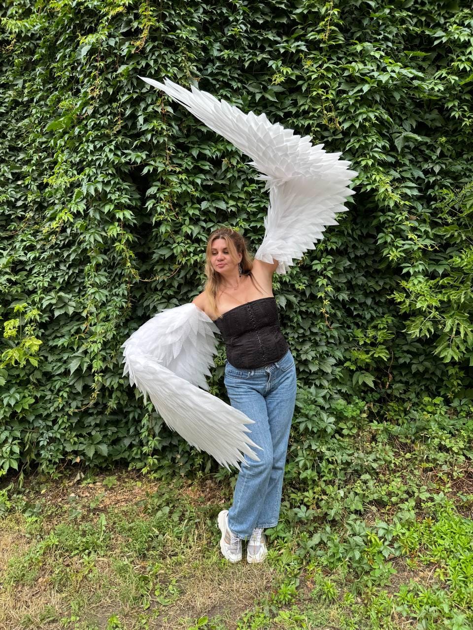 May include: Large, white, articulated feathered wings attached to a black strapless corset top and blue denim jeans. The wings feature layered, pointed white feathers that extend outward, creating a wide, expansive span across the upper body.
