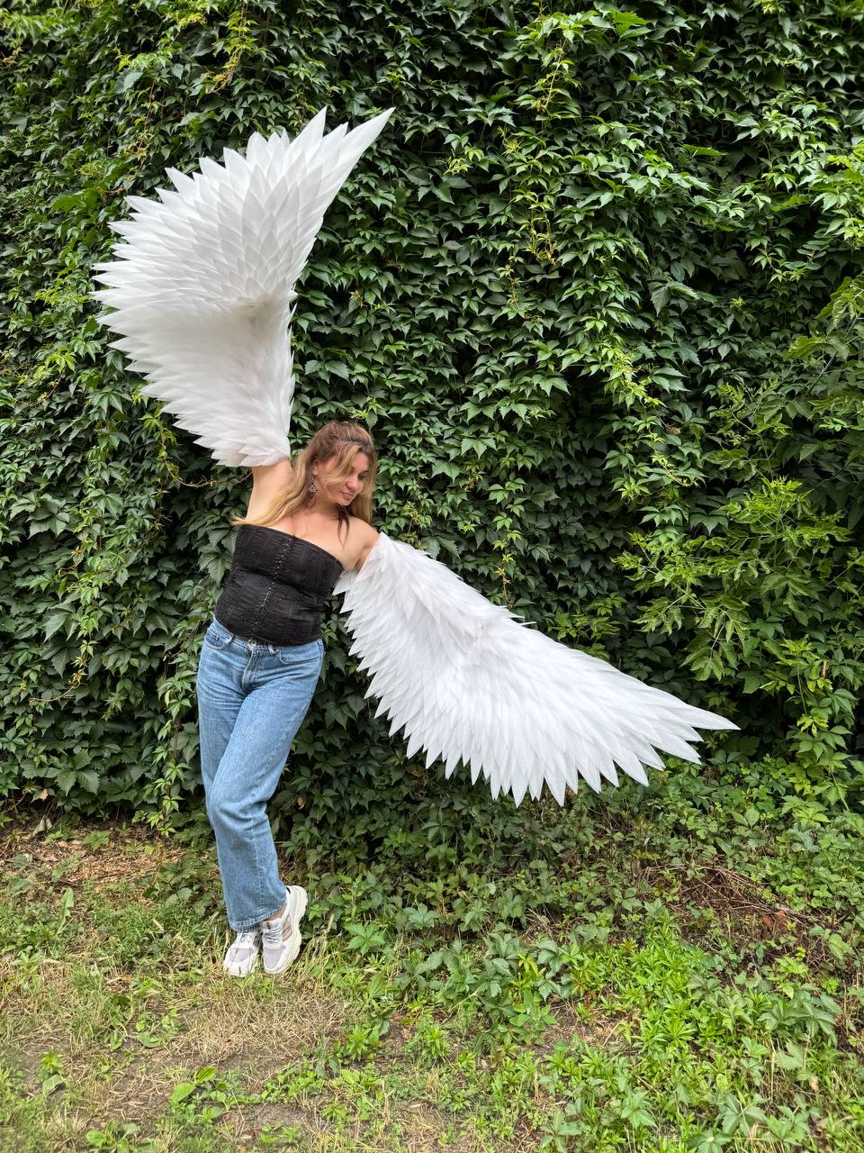 May include: Large white feathered wings attached to a black strapless corset top, blue denim jeans, and white sneakers. The wings feature layered, pointed white feathers extending outward, creating a wide, expansive span across the upper body.