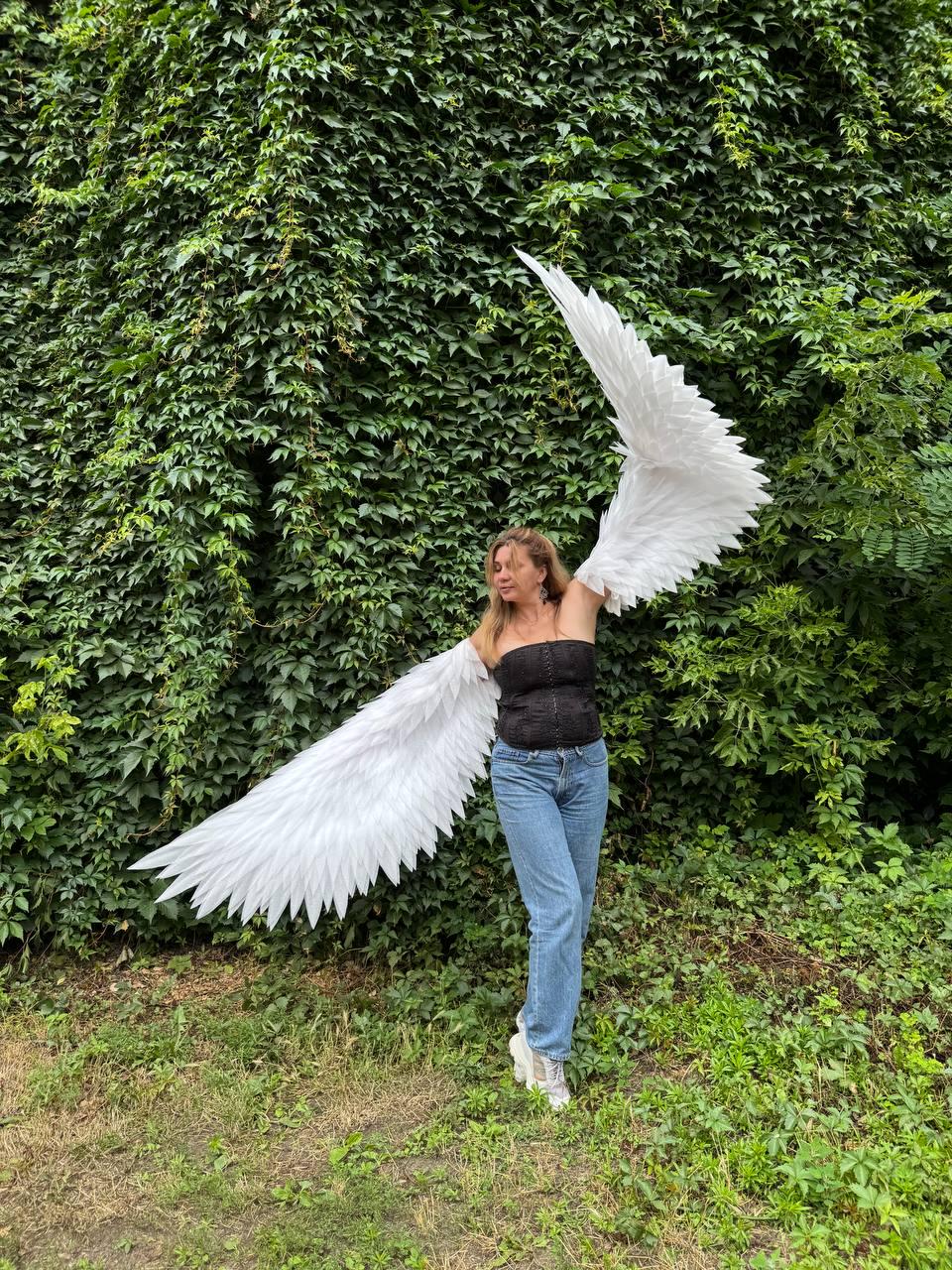 May include: Large, white, feathered-style wings attached to a black strapless corset top and blue denim jeans. The wings are expansive, with one wing extending upward and the other extending downward, featuring layered, pointed white plumage.