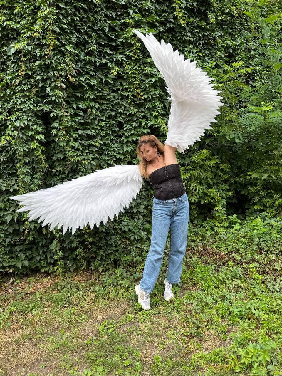 May include: Large white feathered wings attached to a black strapless corset top, paired with light blue denim jeans and white sneakers. The wings feature layered, pointed white feathers extending outward in a wide, expansive span.