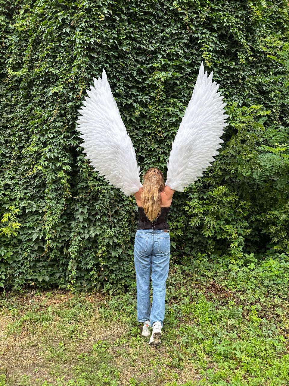 May include: Large white feathered wings attached to a black corset top, worn with light blue denim jeans and white sneakers. The wings feature layered, pointed white feathers extending upward and outward, creating a wide, expansive span.