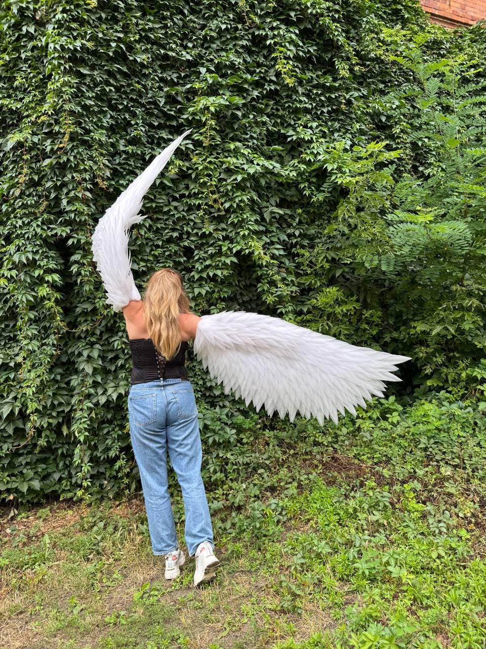 May include: Large white feathered wings attached to a black corset top and light blue denim jeans. The wings are expansive, with one side reaching upward and the other extending horizontally, featuring layered white plumage throughout.