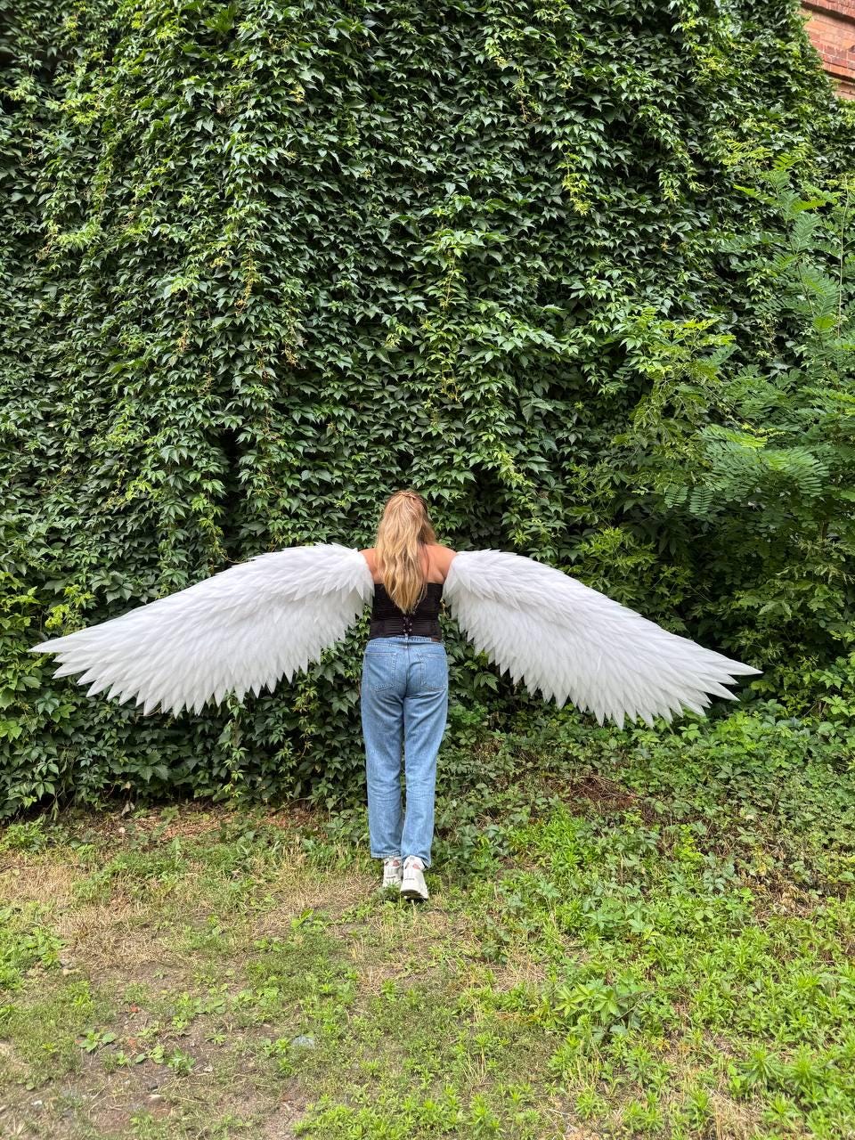 May include: A pair of large, white, feathered angel wings attached to a black corset top worn with light blue denim jeans. The wings extend horizontally from the back, featuring layered, pointed white feathers that span a wide distance.