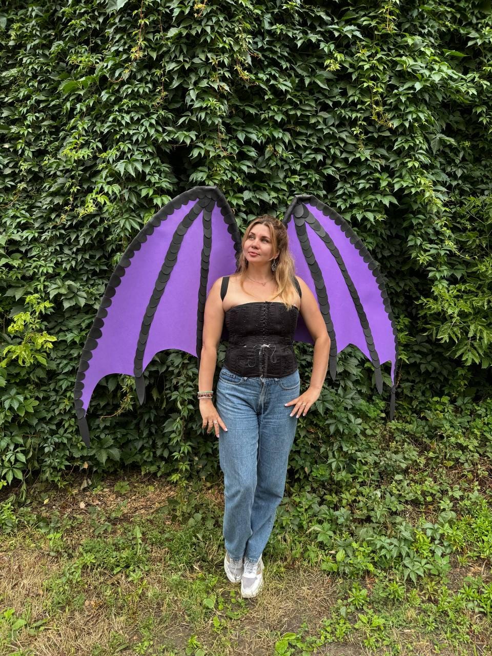 May include: Large purple dragon wings with black scalloped edges and structural ribbing, worn with a black strapless corset top featuring front lacing and blue denim jeans. The wings are expansive and feature a distinct bat-like wing shape.