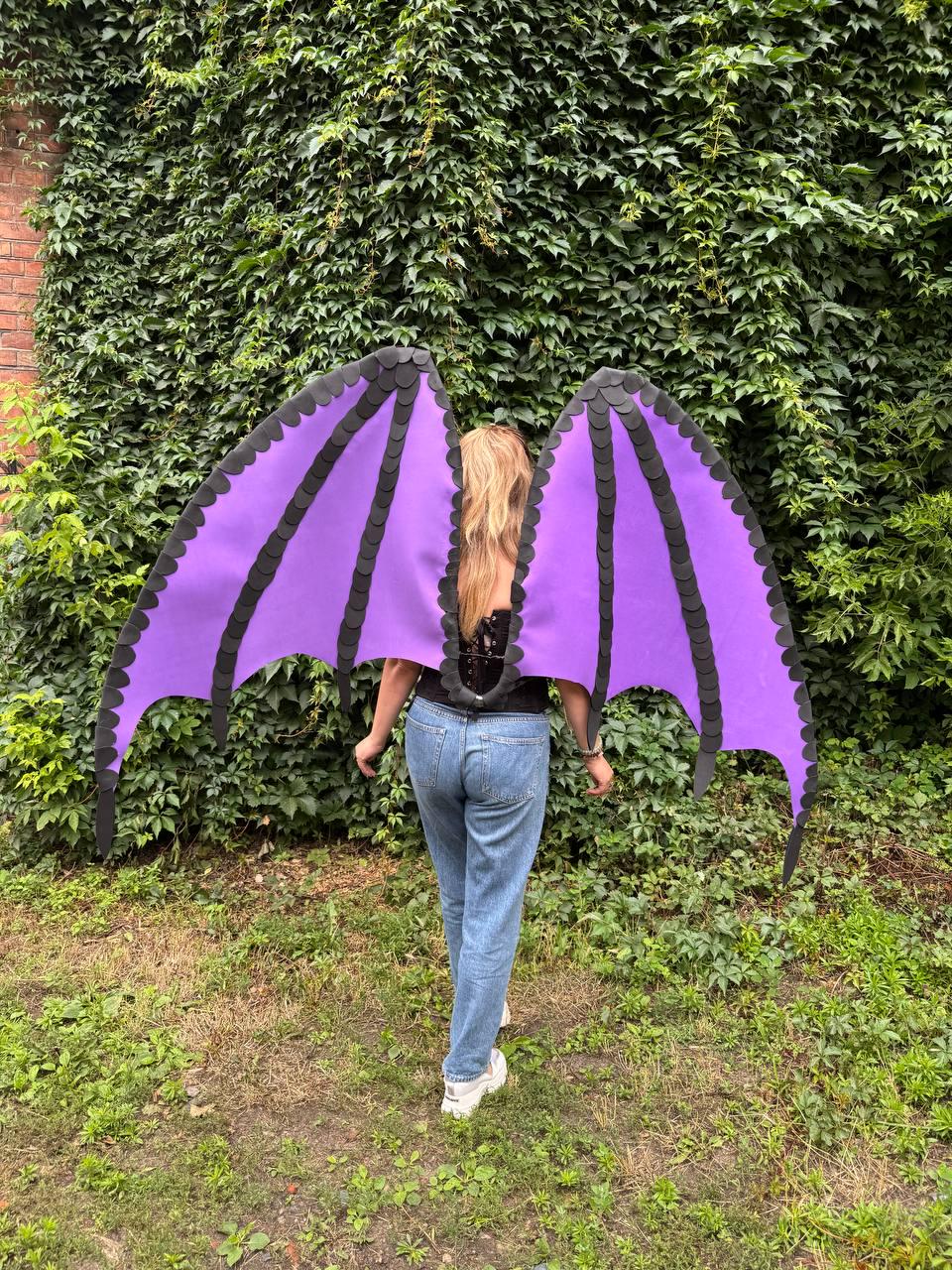 May include: Large dragon-style wings featuring a vibrant purple fabric base with black scalloped edges and structural ribs. The wings are designed to be worn on the back, showcasing a wide, pointed span with detailed layered segments in black.