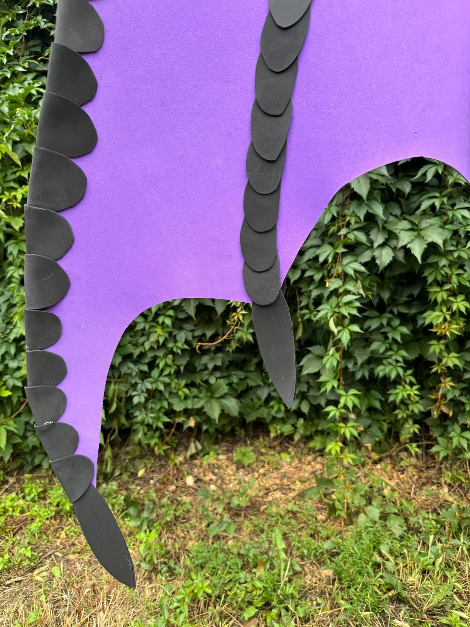 May include: A purple foam costume accessory featuring two vertical rows of overlapping black scale-like cutouts attached to the surface. The item has a curved, irregular shape with pointed ends, designed to resemble a creature's wing or tail structure.