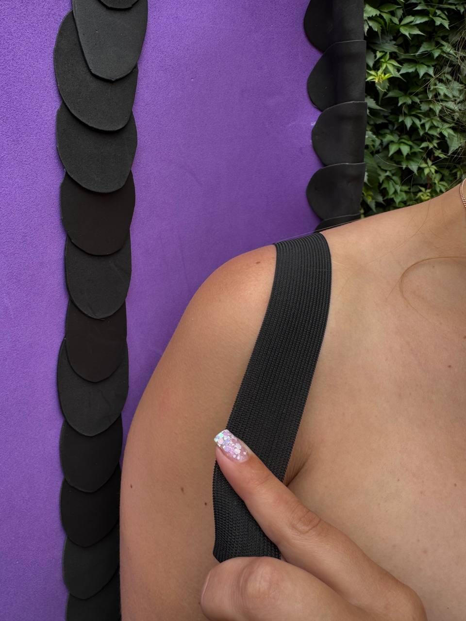 May include: A black elastic strap rests on a shoulder, positioned next to a vibrant purple fabric panel decorated with a vertical row of overlapping, rounded black scales. The design features a clean, contrasting aesthetic with distinct textures.