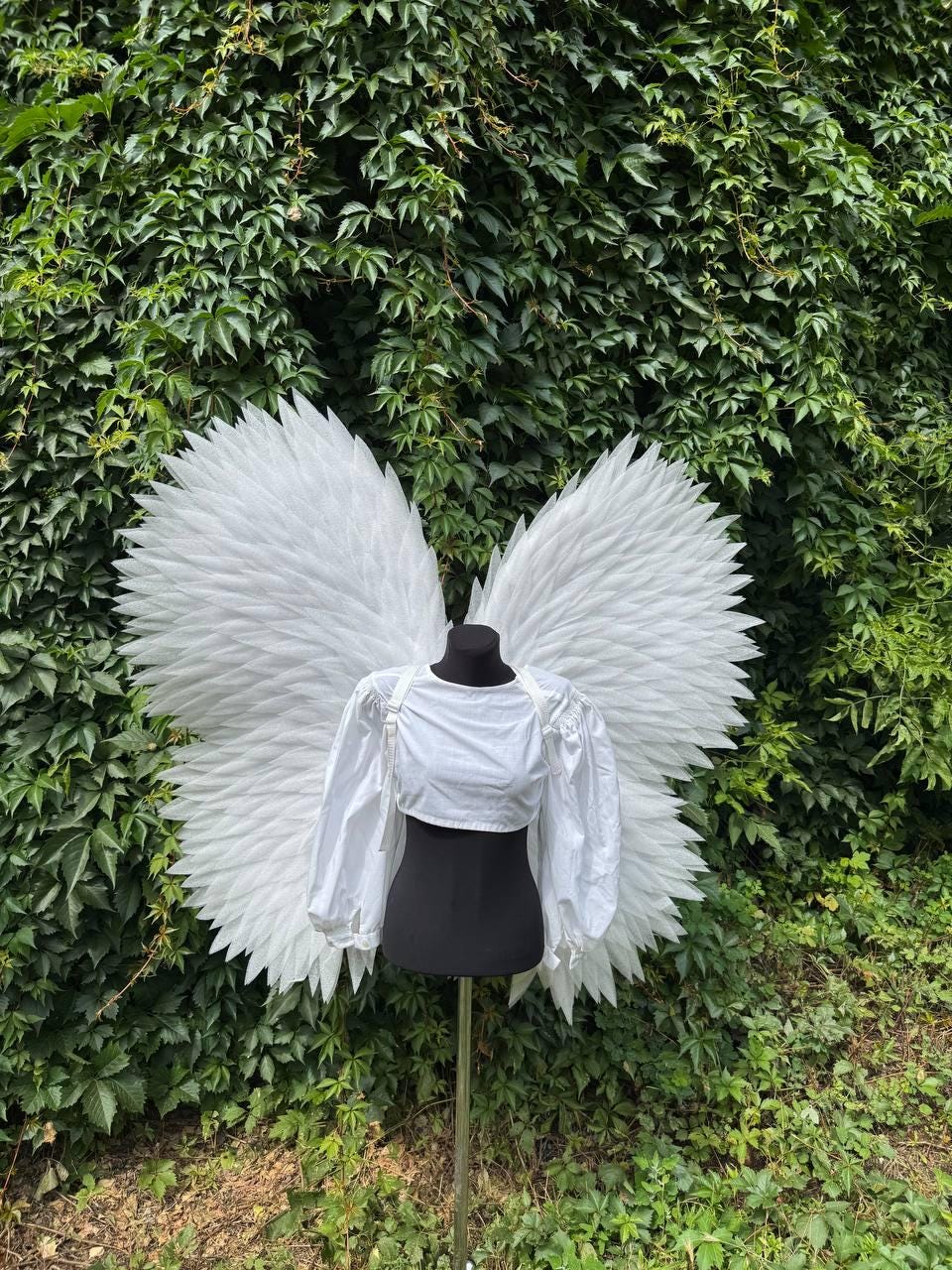 May include: A pair of large, white, feathered angel wings attached to a black mannequin torso. The mannequin is wearing a white, long-sleeved, cropped blouse with a round neckline and gathered cuffs, positioned on a metal stand.
