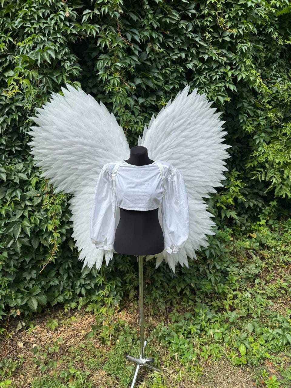 May include: A white crop top with long puff sleeves and large, feathered white angel wings attached to the back. The garment is displayed on a black mannequin stand, featuring a round neckline and adjustable straps for the wings.