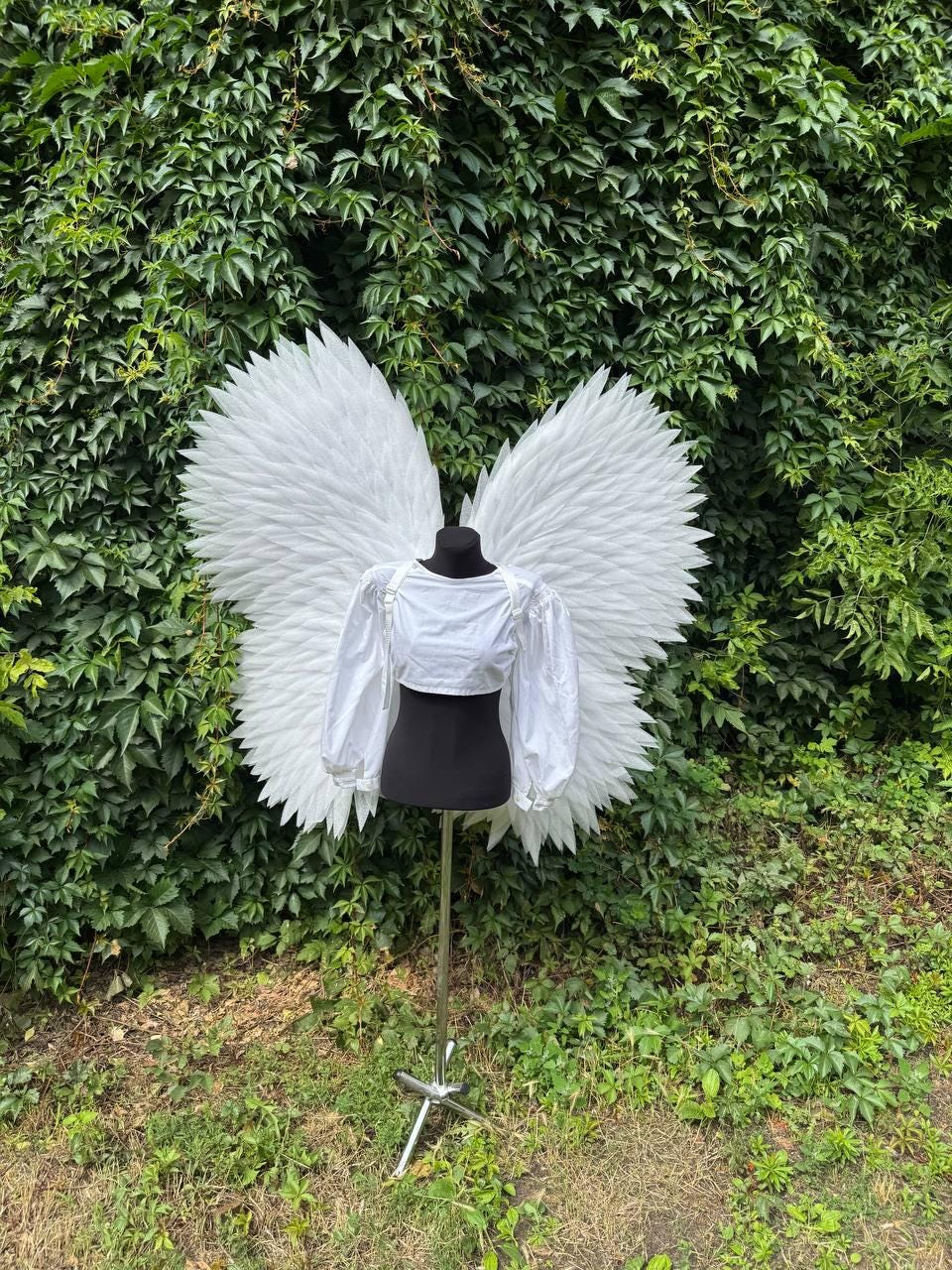 May include: A white cropped long-sleeve top with puffed sleeves displayed on a black mannequin torso. Large, detailed white feathered wings are attached to the back of the mannequin, extending outward to create a wide, expansive wing span.