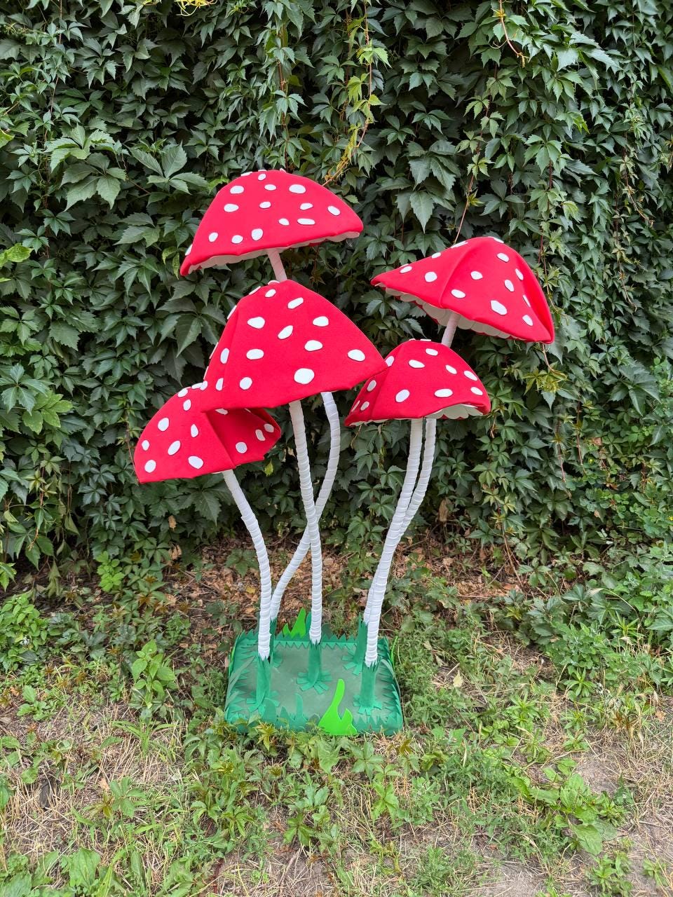 May include: A cluster of five artificial mushrooms with bright red caps featuring white spots. Each mushroom has a slender white stem and is attached to a green rectangular base. The mushrooms vary in height and are arranged in a group.