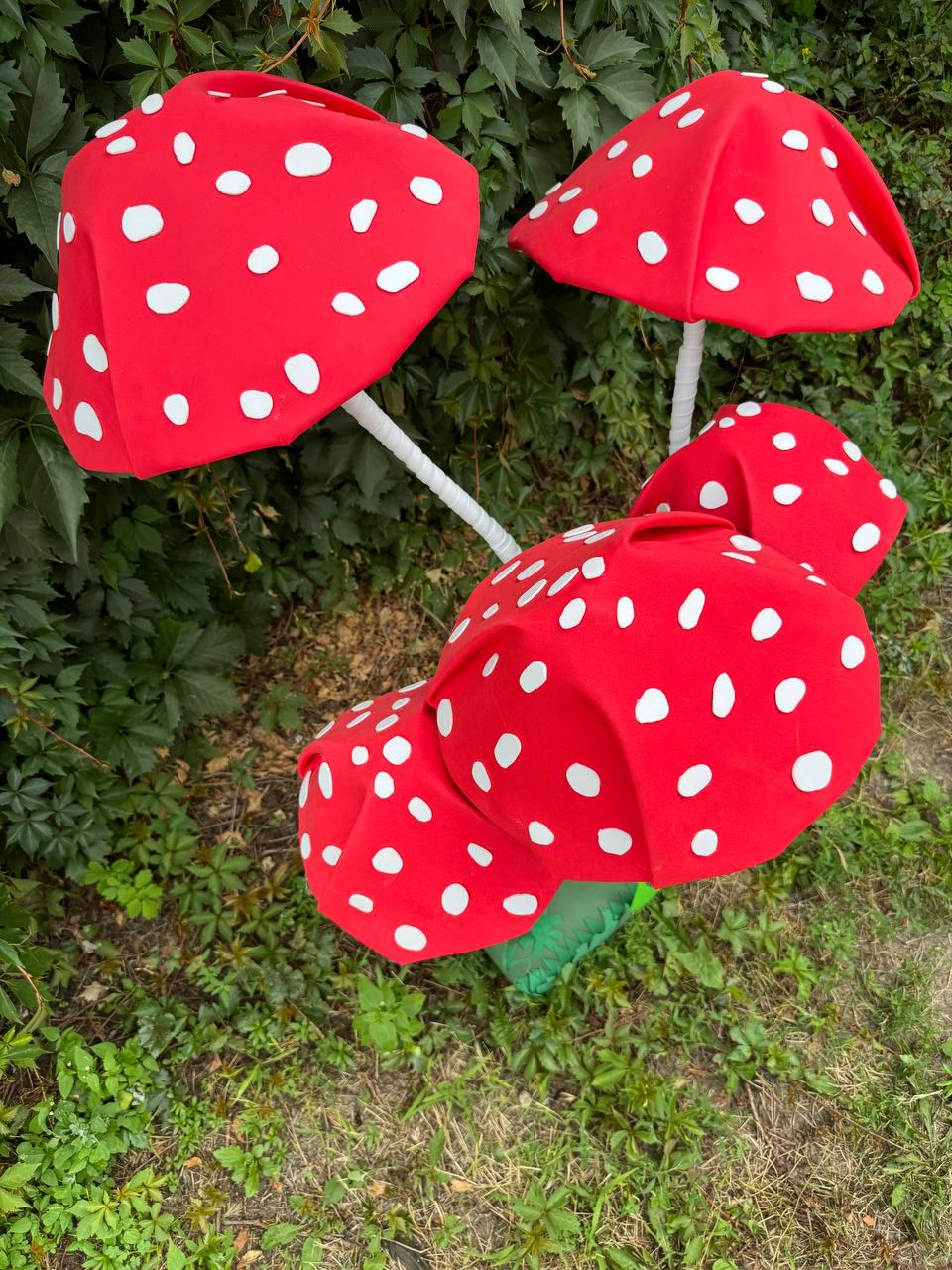 May include: A cluster of four artificial mushroom sculptures featuring bright red rounded caps with scattered white circular spots. Each mushroom has a slender white stem, and the group is arranged at varying heights to create a layered display.