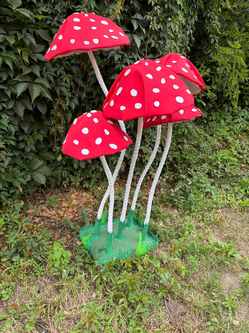May include: A decorative set of four artificial mushrooms featuring bright red caps with white polka dots. Each mushroom has a long, thin, white textured stem and is attached to a green base that mimics grass, creating a cluster of fungi.
