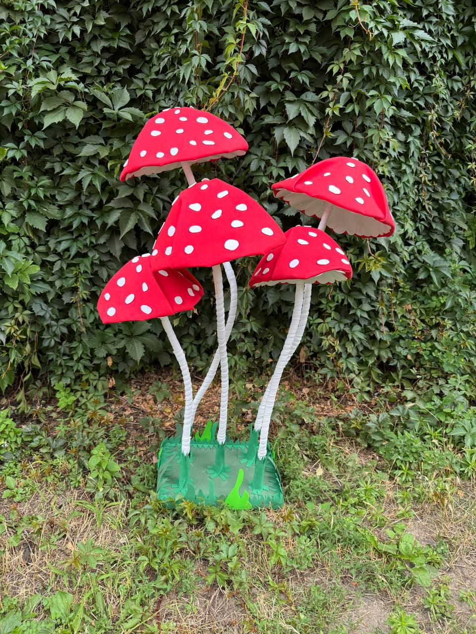 May include: A decorative display featuring four artificial mushrooms with bright red caps and white spots. Each mushroom has a slender, textured white stalk and is attached to a green square base with a cutout grass pattern at the bottom.