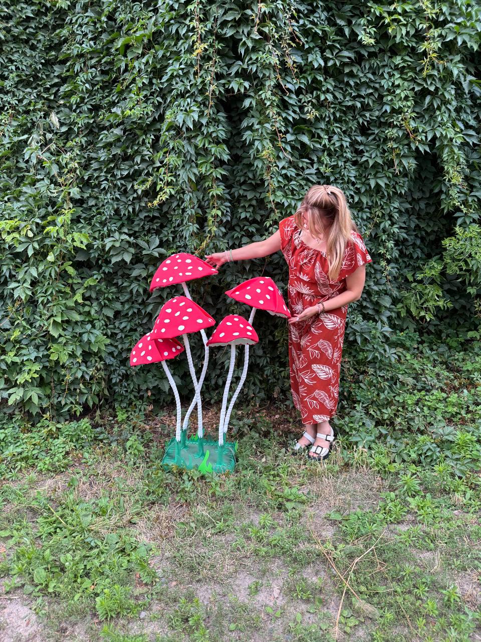 May include: A decorative sculpture featuring four red mushroom caps with white spots, each supported by a thin white stem. The mushrooms are attached to a green rectangular base, creating a cluster of artificial fungi standing upright.