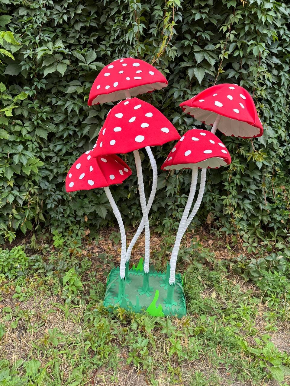 May include: A decorative display featuring four red mushroom caps with white spots, each supported by a thin white stem. The stems are attached to a green square base with jagged edges, creating a cluster of artificial fungi standing upright.