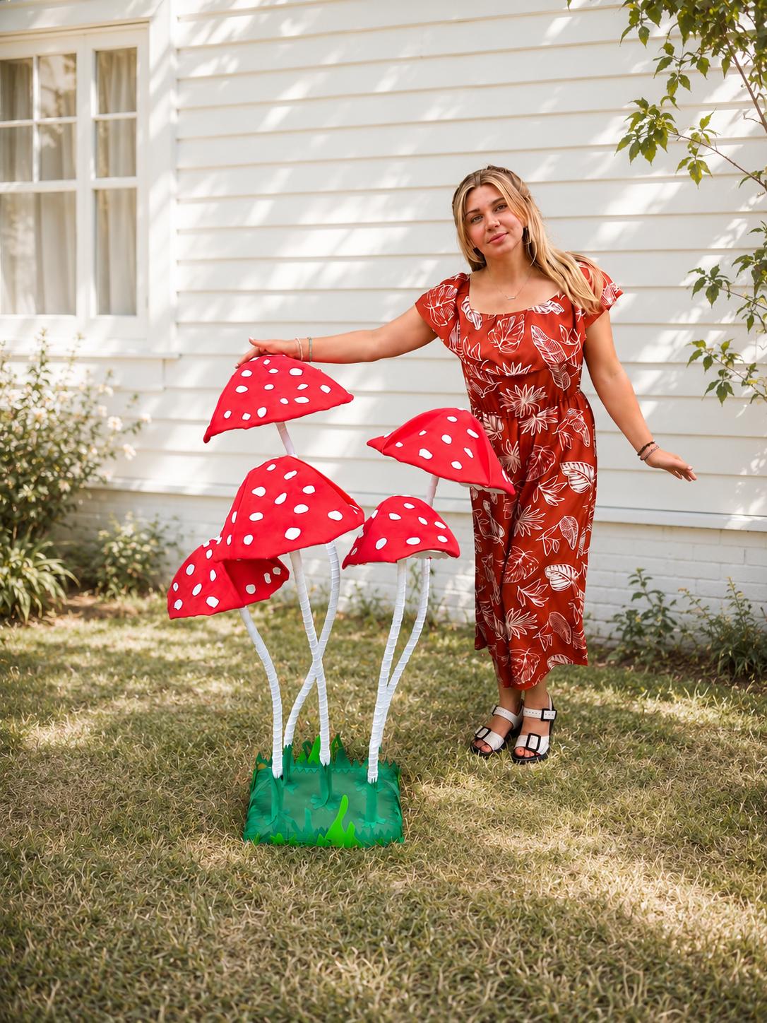 May include: A decorative display featuring four large, red mushroom caps with white polka dots, supported by tall, white, textured stems. The mushrooms are anchored into a square, green base with jagged, grass-like edges at the bottom.