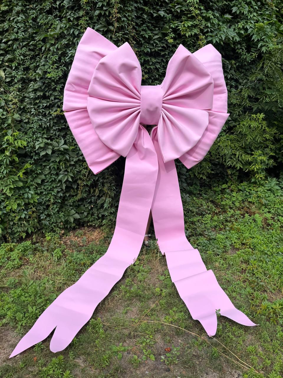 May include: A large, oversized decorative bow in a solid light pink color. The bow features two wide, pleated loops and two long, trailing ribbon tails that extend downward, creating a prominent and structured display piece with a smooth finish.