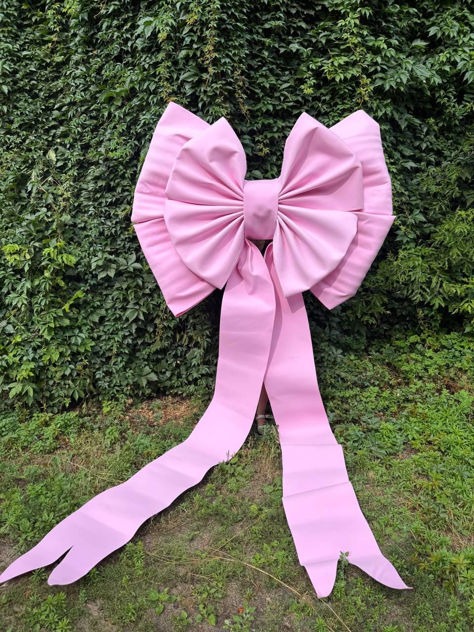May include: A large, oversized light pink bow constructed from a smooth, matte material. The bow features two wide, voluminous loops and two long, trailing ribbon tails that extend downward, creating a prominent and structured decorative shape.