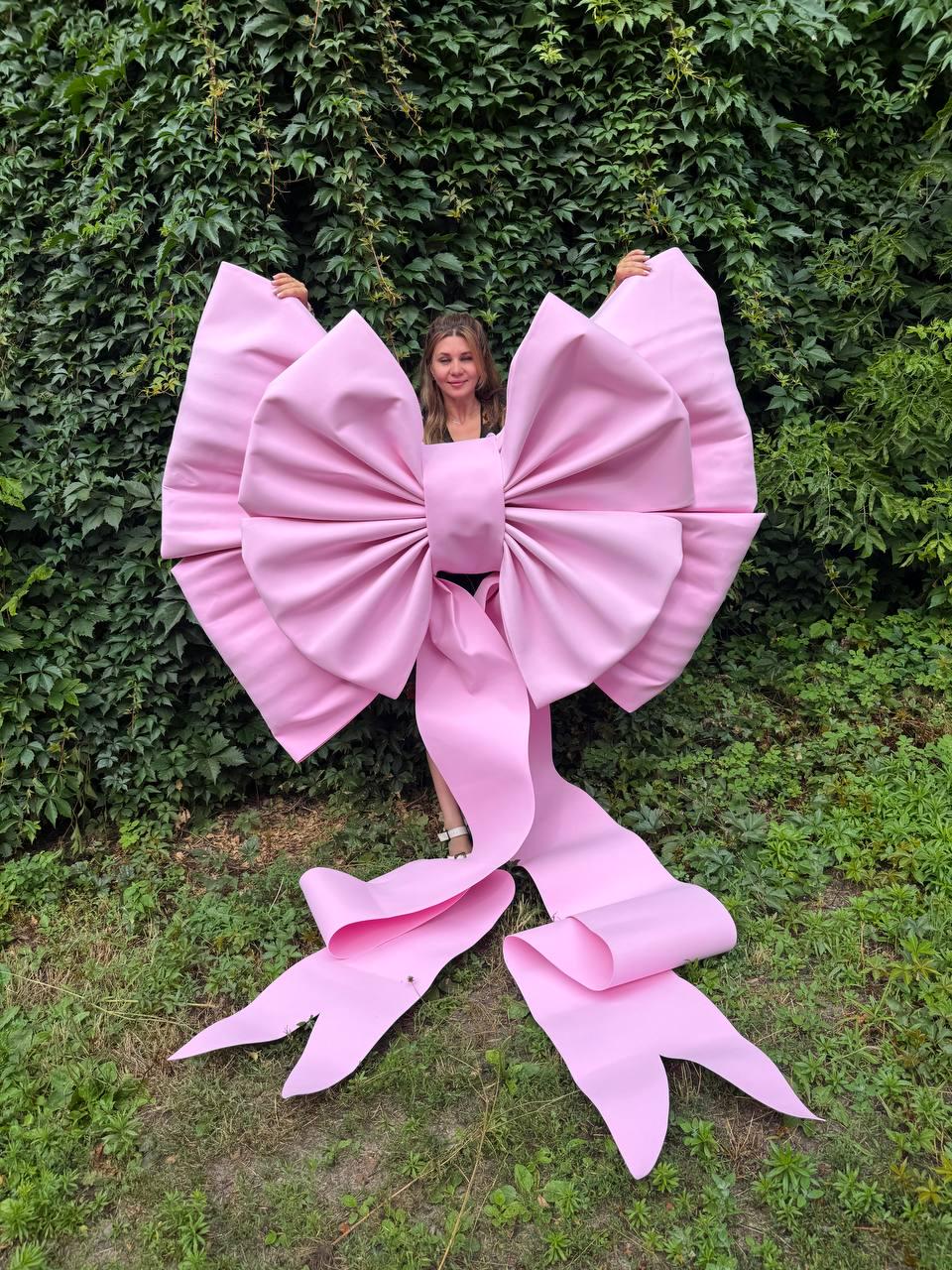 May include: An oversized, light pink decorative bow made of smooth material. The bow features large, voluminous loops and two long, flowing ribbon tails that extend downward, creating a prominent and substantial display piece.