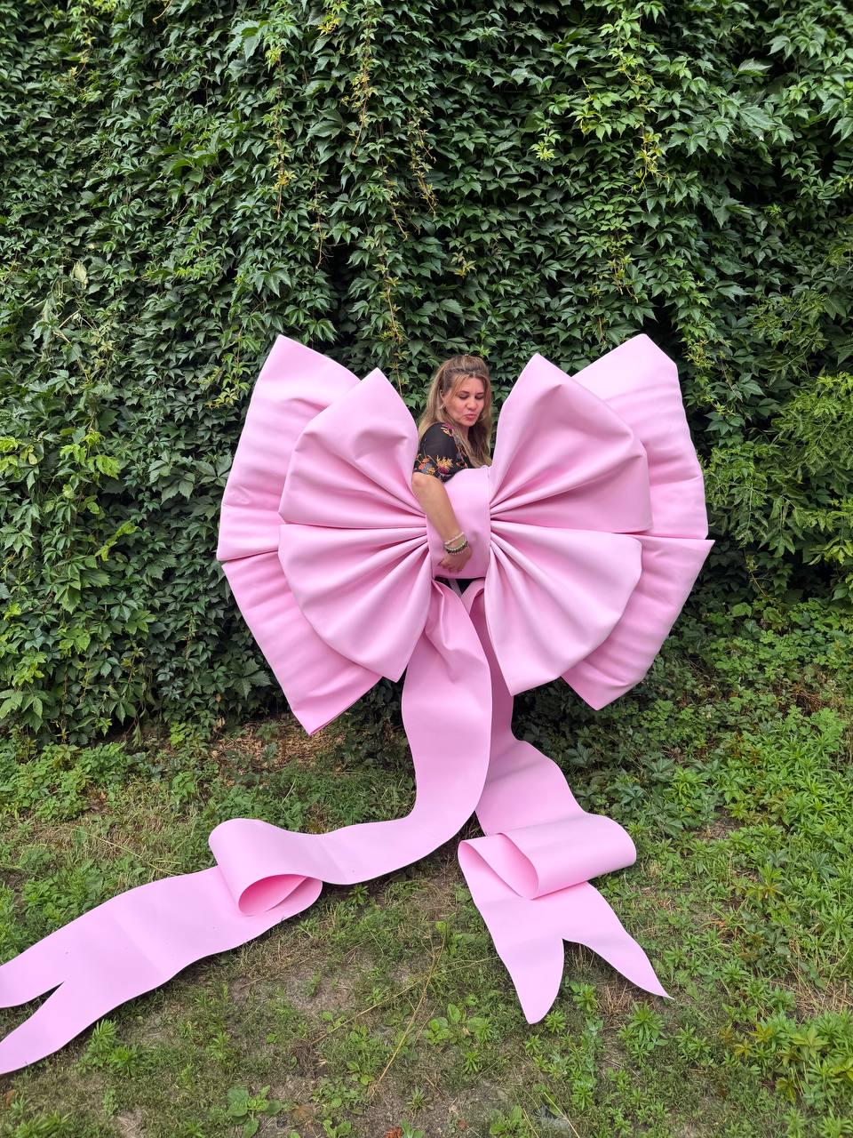 May include: An oversized, light pink decorative bow constructed from a smooth, matte material. The bow features large, voluminous loops and two long, trailing ribbon tails that extend downward, creating a prominent and sculptural display piece.