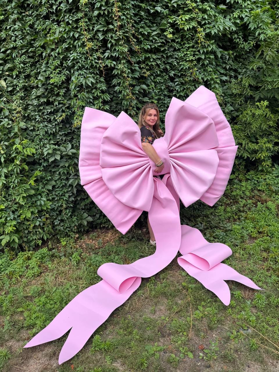 May include: An oversized, light pink decorative bow constructed from a smooth, matte material. The bow features large, voluminous loops and two long, trailing ribbon tails that extend downward, creating a dramatic and substantial display piece.
