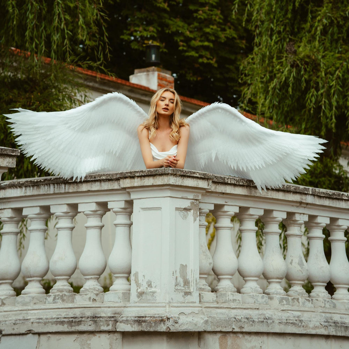May include: A pair of large, white, feathered angel wings attached to a white garment. The wings feature layered, pointed plumage extending horizontally, creating a wide span. The design is structured with individual feather-like textures.