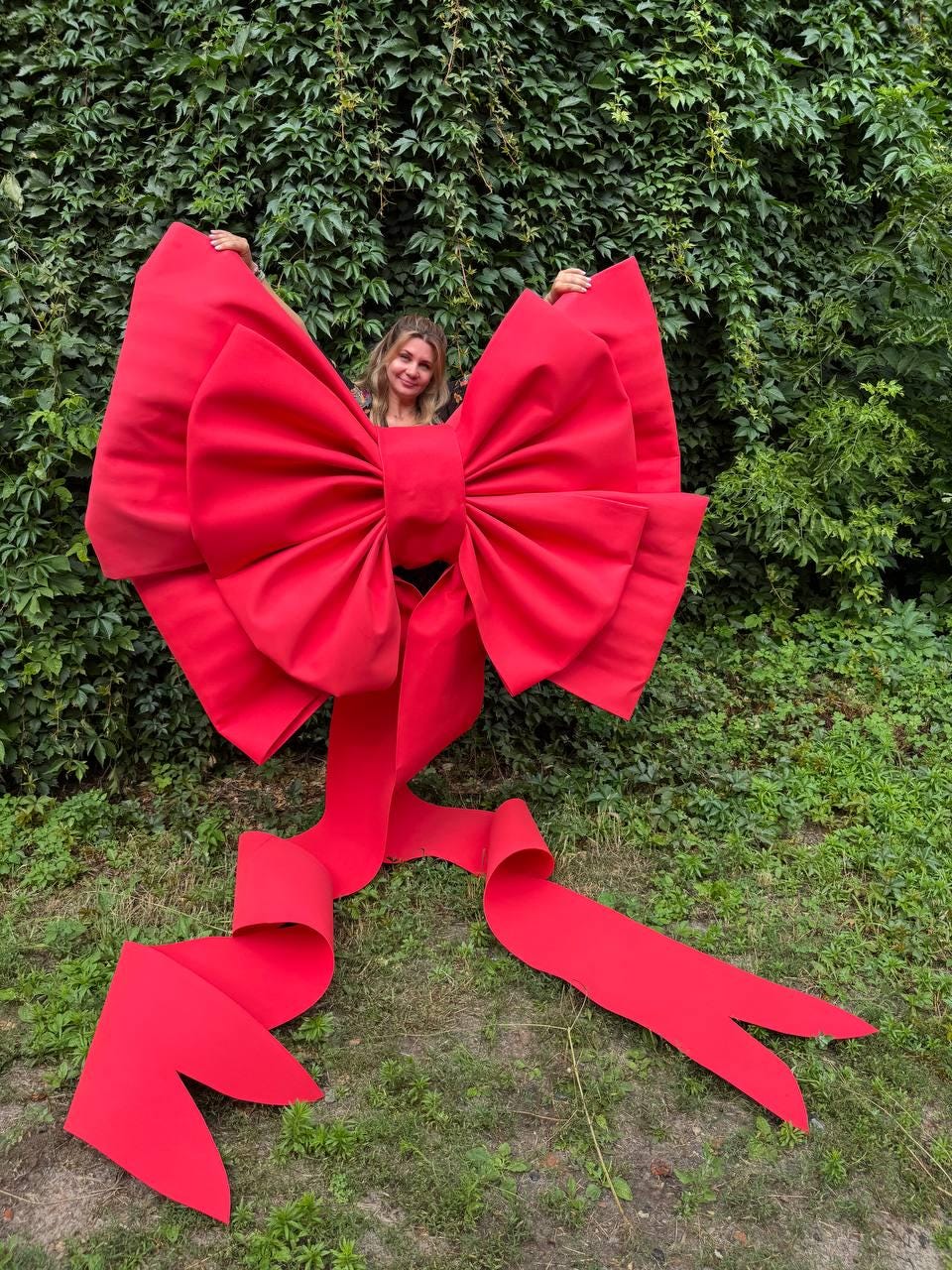 May include: An oversized, bright red decorative bow constructed with stiff, structured fabric. The bow features two large, voluminous loops and two long, trailing ribbon tails that extend downward, each ending in a V-shaped notch at the bottom.