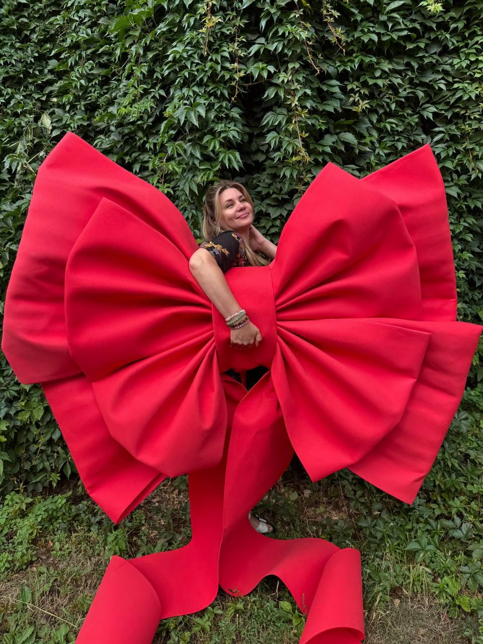 May include: An oversized, bright red decorative bow constructed from a smooth, matte material. The bow features large, voluminous loops and two long, trailing ribbon ends that extend downward, creating a bold and striking visual statement piece.