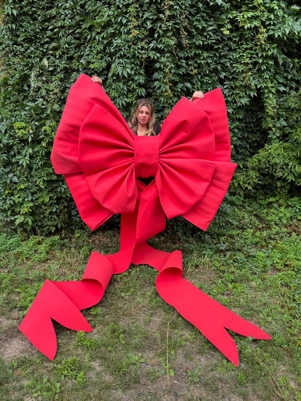 May include: An oversized, bright red decorative bow made of stiff fabric. The bow features two large, voluminous loops with pleated details and two long, wide ribbon tails that extend downward, ending in pointed, V-shaped cutouts at the bottom edges.