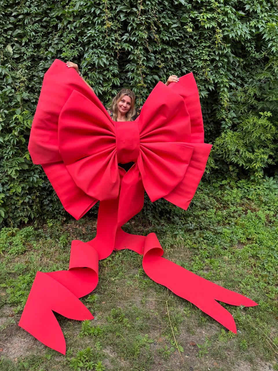 May include: An oversized, bright red decorative bow constructed from stiff material. The bow features two large, voluminous loops and two long, trailing ribbon tails that extend downward, each ending in a V-shaped notch at the bottom edge.
