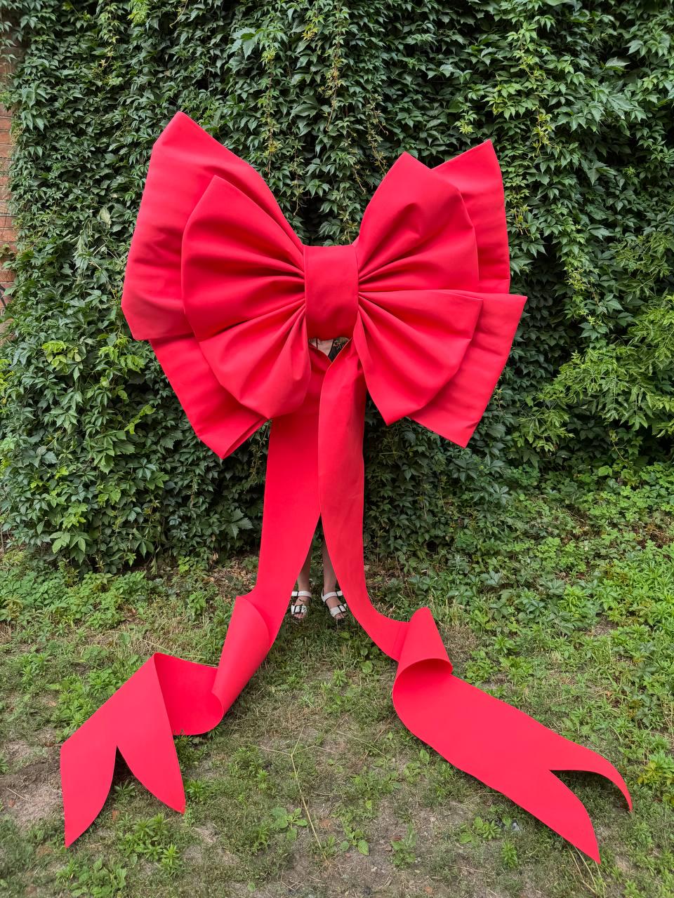 May include: An oversized, vibrant red decorative bow constructed from stiff fabric. The bow features two large, voluminous loops and two long, trailing ribbon tails that extend downward, ending in pointed, V-shaped cutouts at the bottom edges.