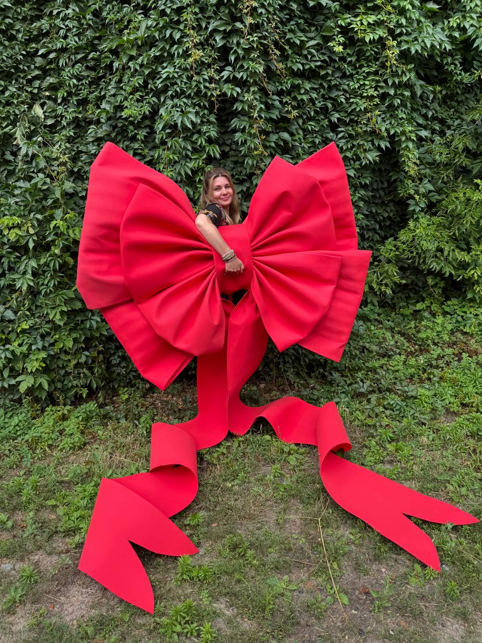 May include: An oversized, bright red decorative bow constructed from stiff material. The bow features two large, voluminous loops and two long, trailing ribbon tails that extend downward, creating a dramatic and bold visual statement piece.