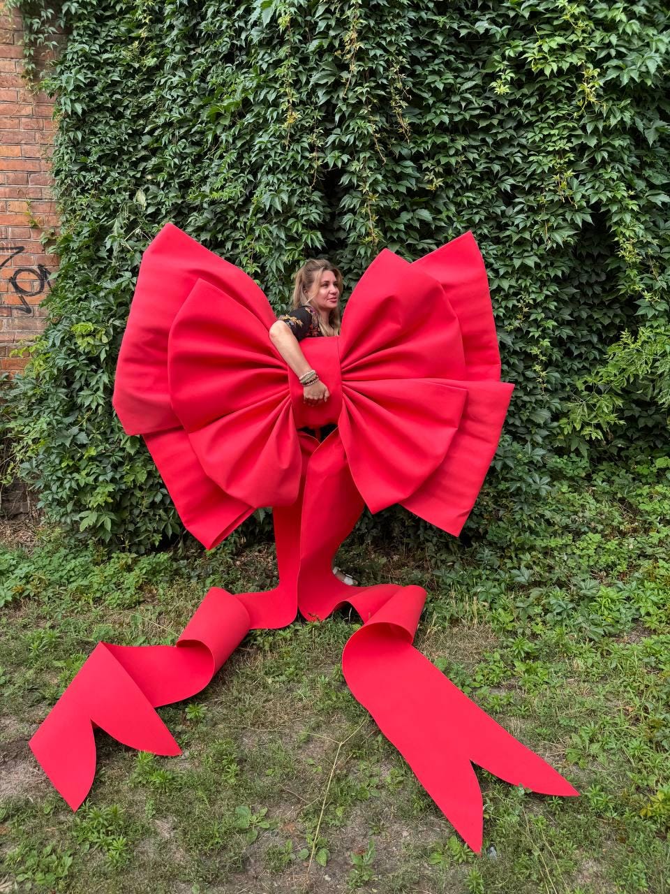 May include: An oversized, bright red decorative bow featuring two large, voluminous loops and two long, trailing ribbon tails with notched ends. The bow is constructed from a smooth, solid red material and is designed to be held or displayed.