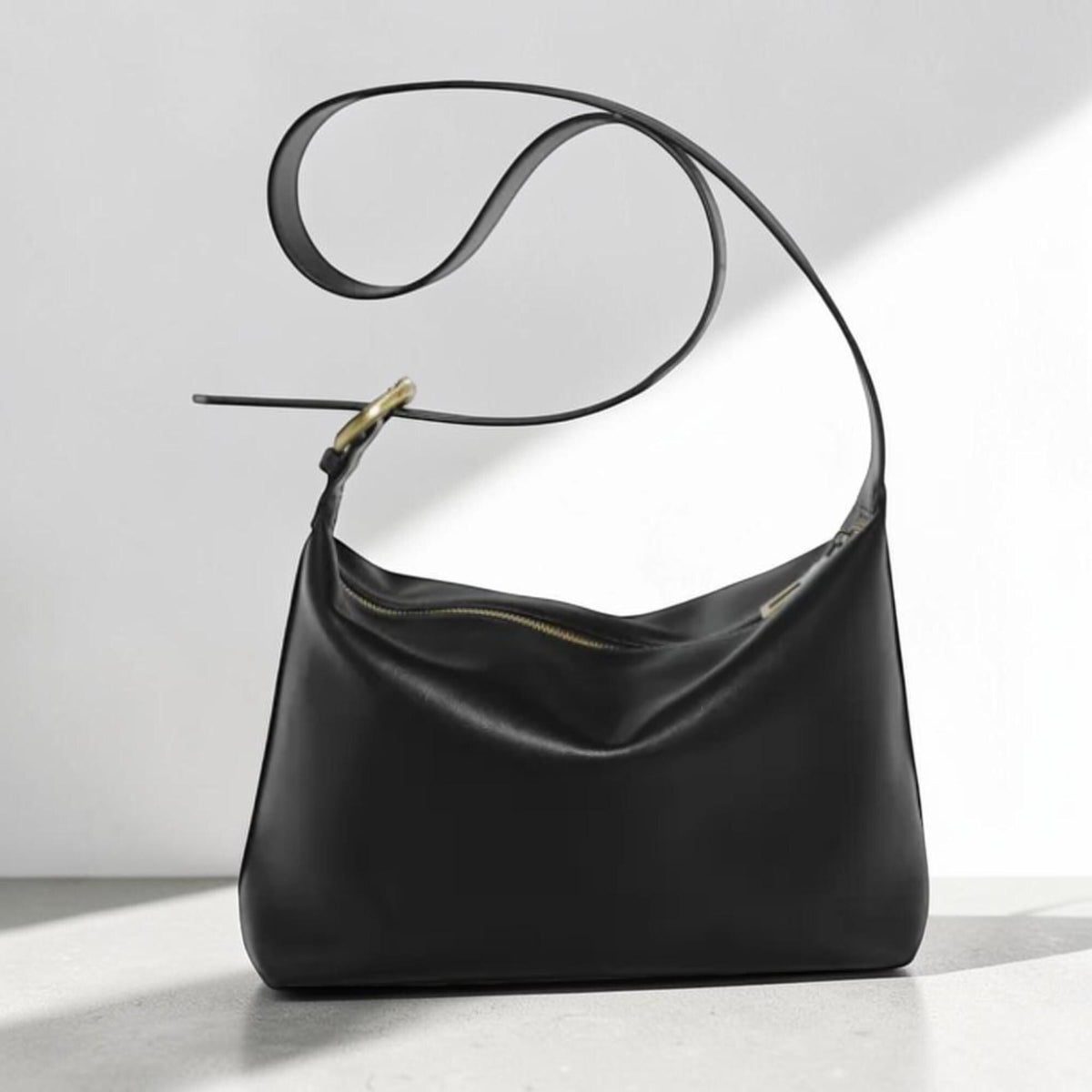 May include: A smooth black leather shoulder bag with a soft, slouchy silhouette. The bag features a single long strap with a gold-toned metal buckle adjustment and a top zipper closure with a gold-toned pull, creating a clean and simple aesthetic.