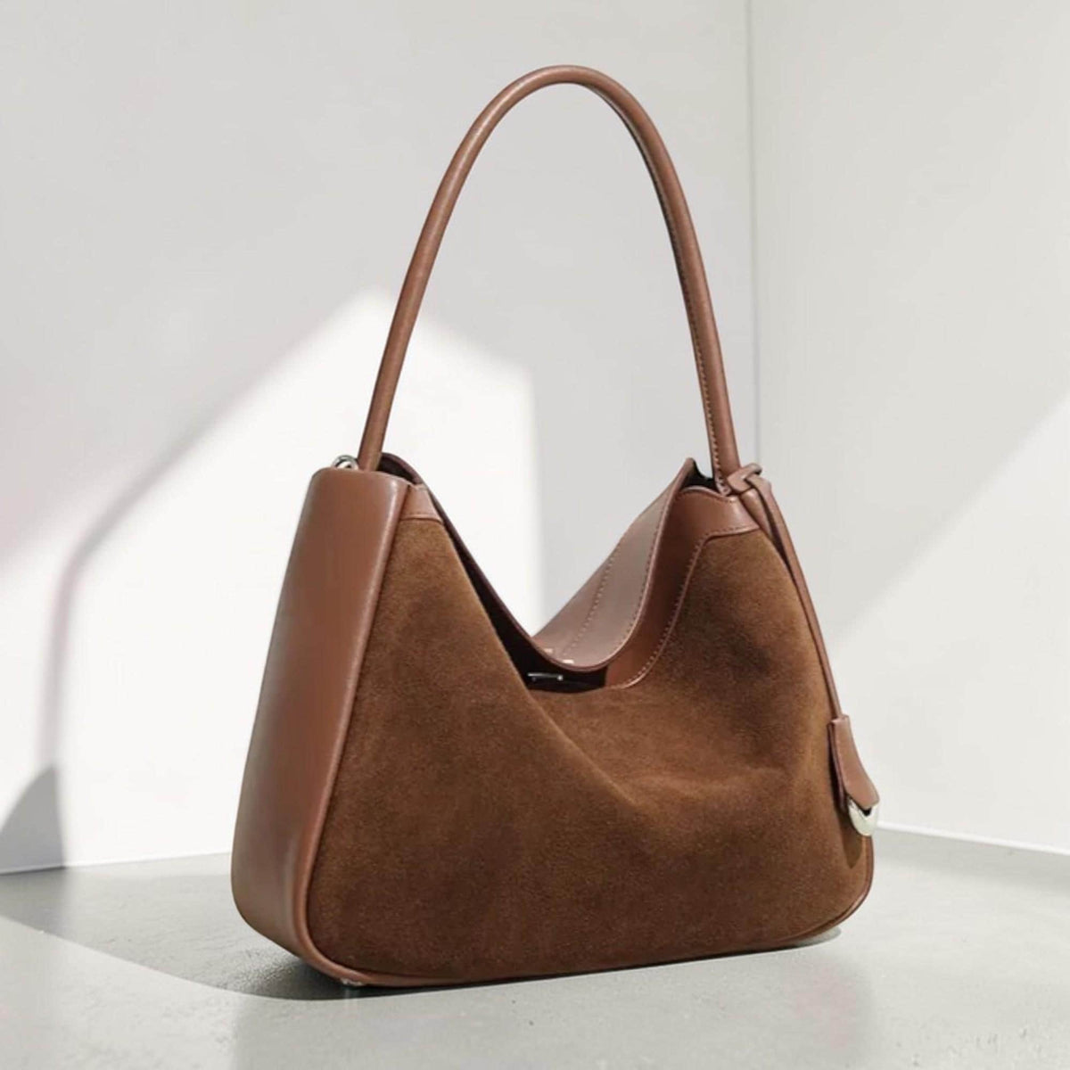 May include: A brown suede shoulder bag with a curved handle. The bag features contrasting leather trim and a simple, elegant design. The bag's dimensions are approximately 30 cm wide and 25 cm high.