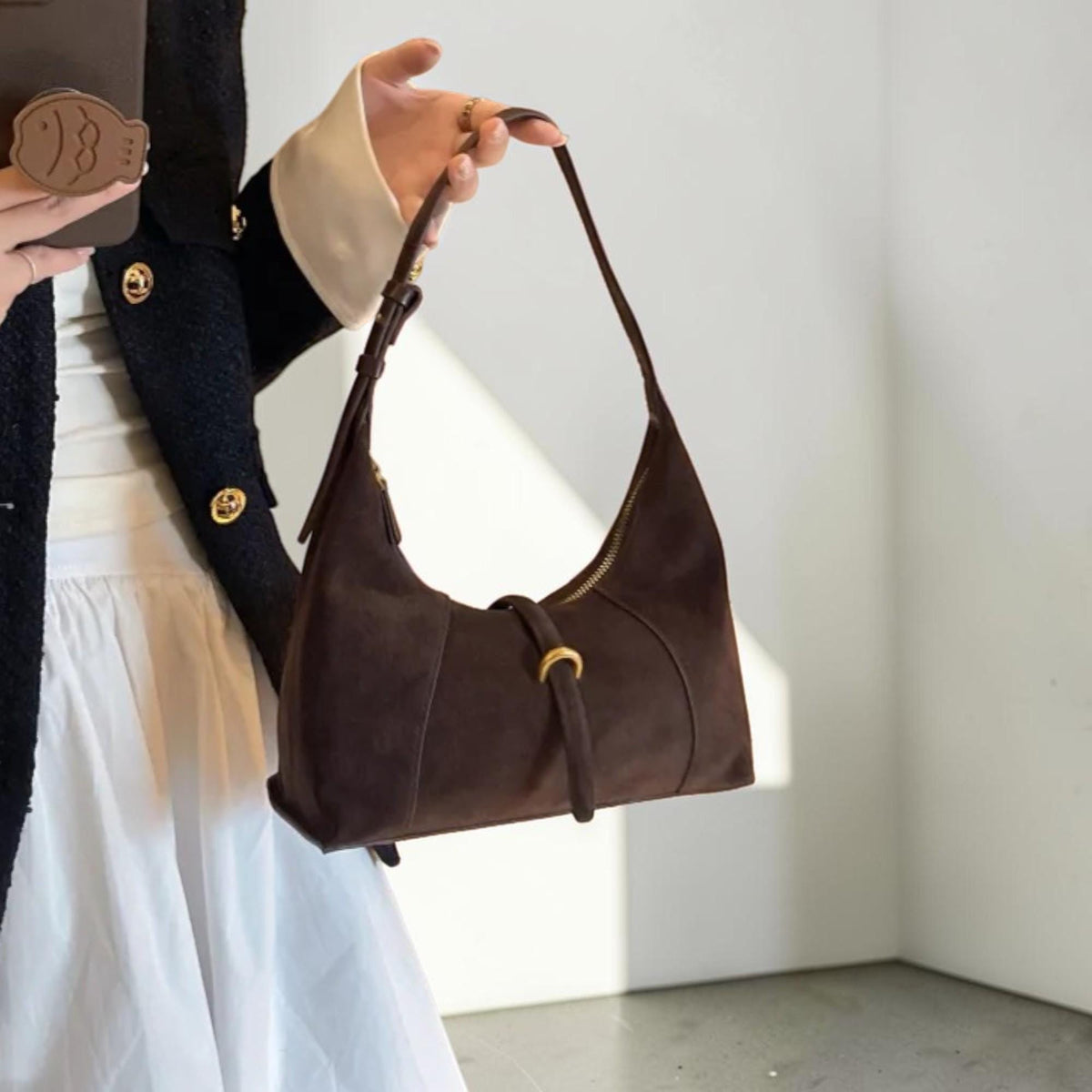 May include: A dark brown suede shoulder bag with a curved shape and a long strap. The bag features a gold-coloured zip and a decorative buckle with a gold ring. The bag is held by a person.