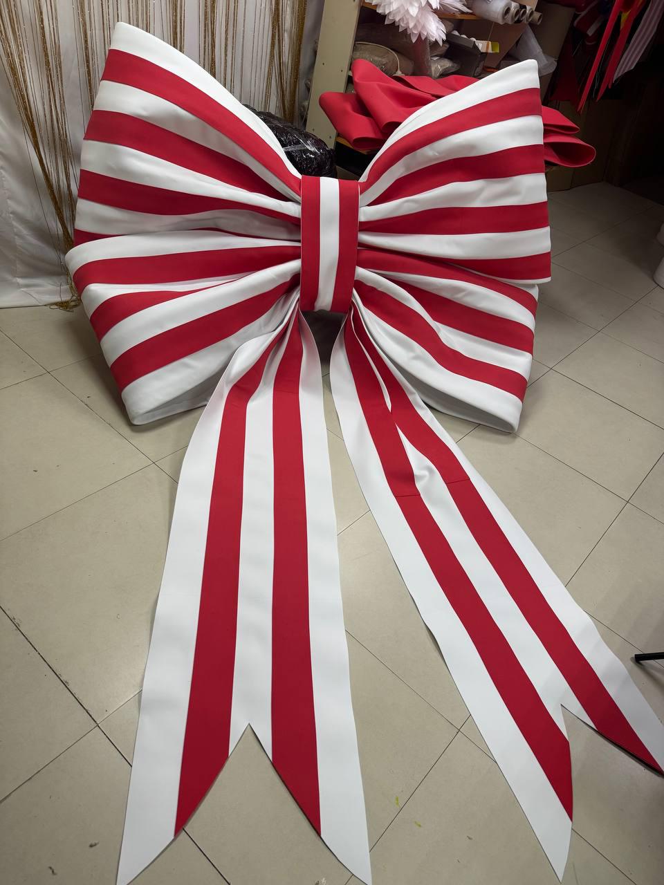 May include: A large decorative bow featuring a repeating pattern of vertical red and white stripes. The bow has two wide, voluminous loops and two long, dangling ribbon tails that end in pointed, V-shaped notches at the bottom edges.