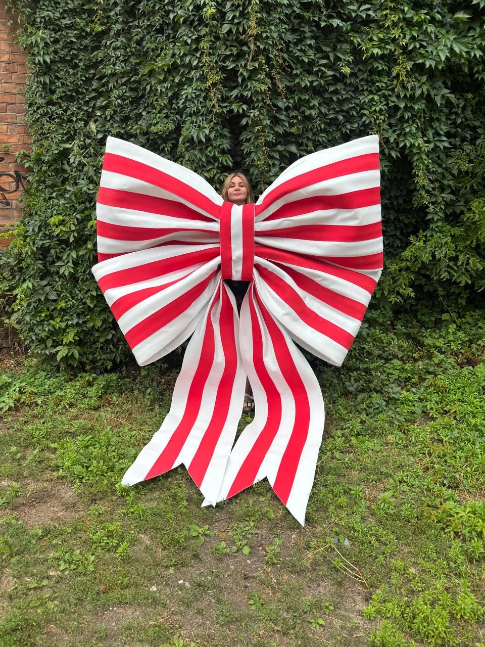 May include: An oversized decorative bow featuring a bold pattern of alternating horizontal and vertical red and white stripes. The large bow is constructed with wide fabric panels that form two expansive loops and two long, trailing ribbon ends.