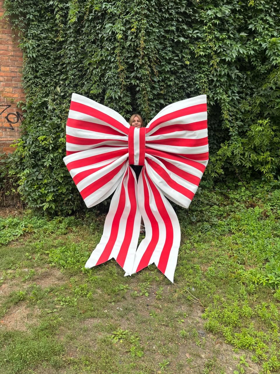 May include: An oversized decorative bow featuring a repeating pattern of bold red and white vertical stripes. The bow has two large, voluminous loops and two long, wide tails that extend downward, creating a prominent and structured display piece.