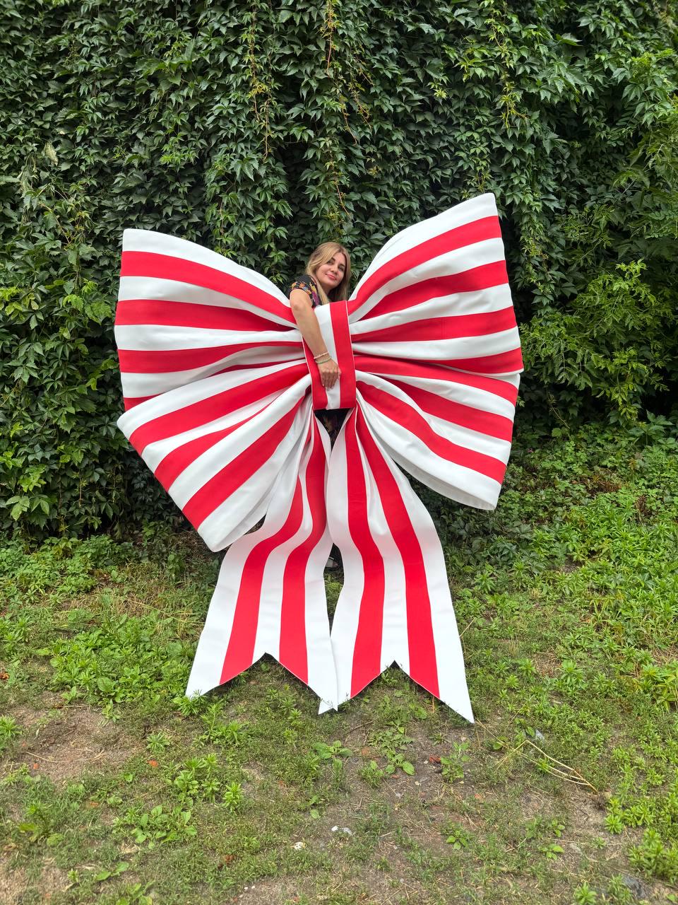 May include: An oversized decorative bow featuring bold vertical red and white stripes. The large, structured bow has two wide loops and two long, pointed tails hanging downward, creating a prominent display piece with a classic striped pattern.