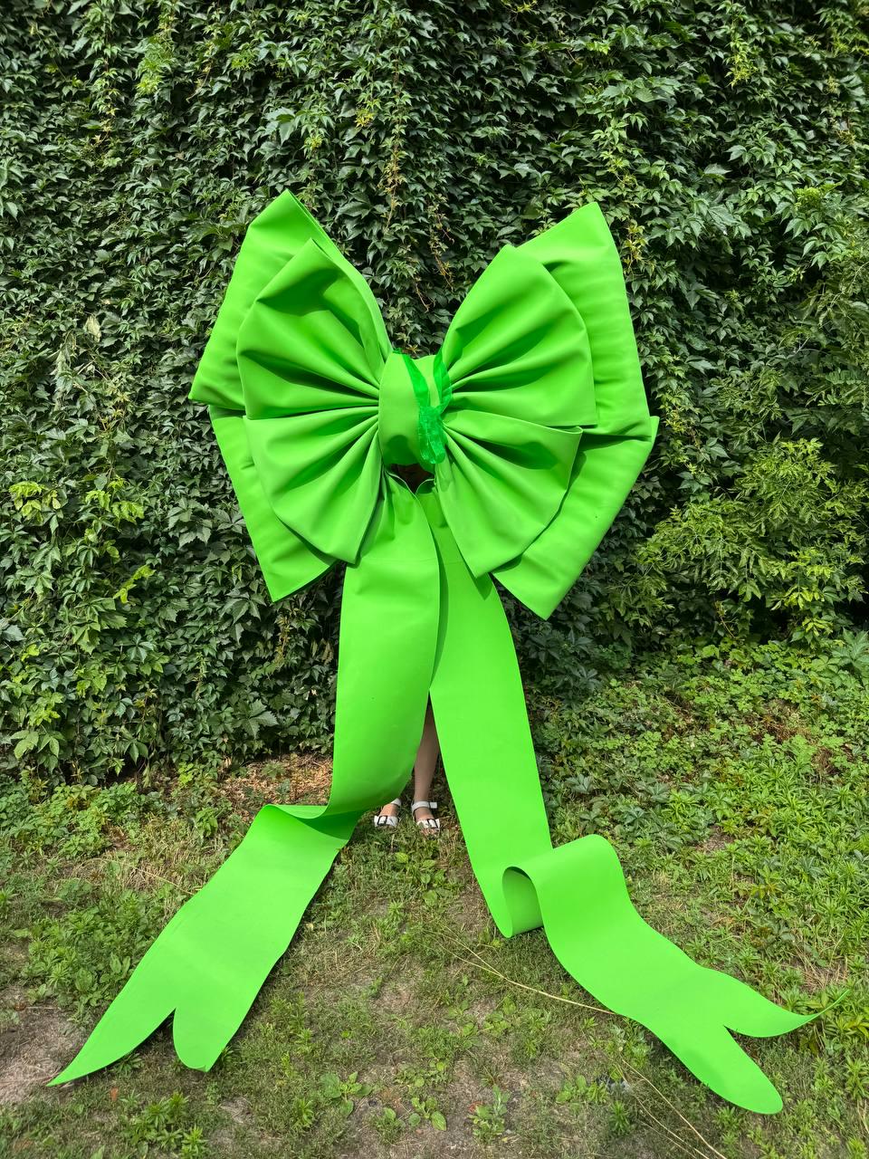 May include: A large, bright lime green decorative bow featuring a central knot and two long, flowing ribbon tails that extend downward. The bow is constructed with multiple structured folds and pleats, creating a voluminous, oversized appearance.