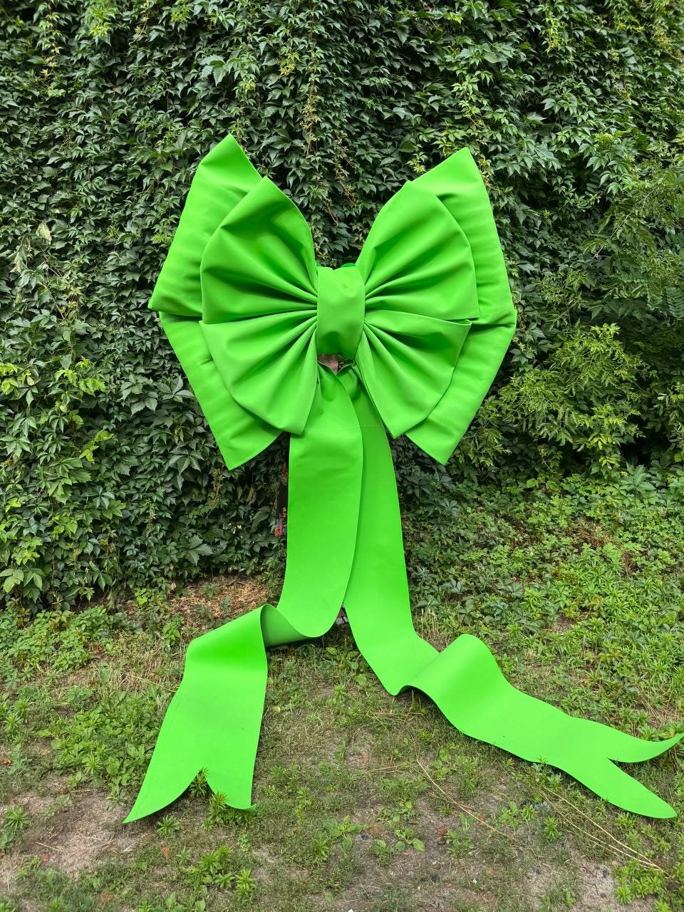 May include: A large, bright lime green decorative bow made of smooth, matte material. The bow features two wide, pleated loops and two long, flowing ribbon tails that extend downward, ending in pointed, V-shaped cutouts at the bottom edges.
