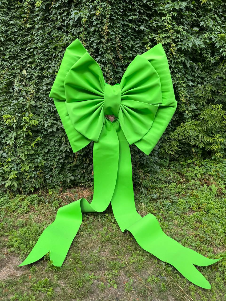 May include: A large, bright lime green decorative bow featuring multiple layered loops and two long, flowing ribbon tails that extend downward. The bow has a smooth, matte finish and a structured, symmetrical design with pointed ends on the tails.